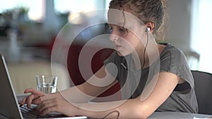 Young caucasian smart schoolgirl with headphones typing in notebook, watching on laptop, smiling and doing homework