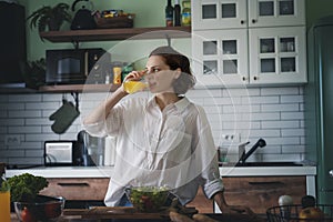 Young caucasian smiling woman drinking orange juice while standing in the kitchen at home