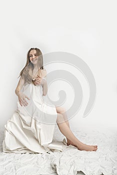 Young caucasian smiling woman is sitting covered herself with white sheet and poses. Concept of good morning. White background.