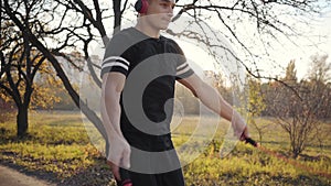 Young Caucasian sportsman in headphones and black sportswear jumping rope in the autumn park on sunset. Athletic boy
