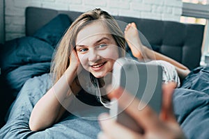 A young Caucasian teenage girl in pajamas lies in bed and holds a phone in front of him and communicates with friends with smile b photo