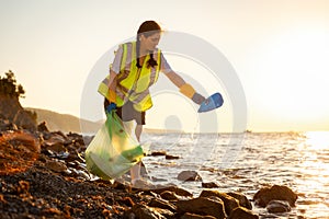 Young Caucasian volunteer woman in rubber gloves collects garbage on wild pebble coast. Copy space. Concept of