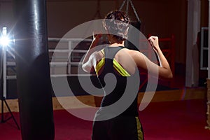 A young Caucasian woman actively trains in the gym in front of a punching bag. The view from the back