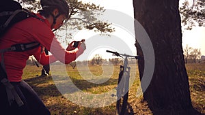 Young caucasian woman athlete tourist cyclist makes photo on phone, photographs his bike standing near tree on nature photo