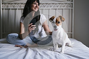 Young caucasian woman on bed using mobile phone. Cute small dog sitting besides. Love for animals and technology concept.