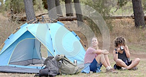 Young Caucasian woman and biracial woman relax beside a blue tent in the woods