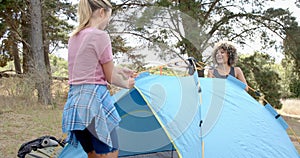 Young Caucasian woman and biracial woman set up a blue tent outdoors