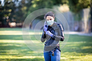 Young Caucasian woman in black leather jacket and surgical mask takes on purple latex gloves