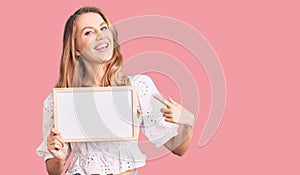 Young caucasian woman with blond hair holding blank empty banner smiling happy pointing with hand and finger