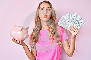 Young caucasian woman with blond hair holding dollars and piggy bank puffing cheeks with funny face