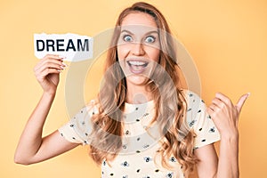 Young caucasian woman with blond hair holding dream word paper pointing thumb up to the side smiling happy with open mouth