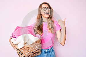 Young caucasian woman with blond hair holding laundry basket pointing thumb up to the side smiling happy with open mouth