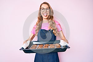 Young caucasian woman with blond hair wearing baker uniform holding homemade cookies sticking tongue out happy with funny