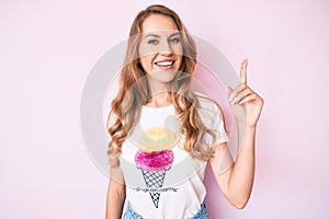 Young caucasian woman with blond hair wearing casual summer clothes surprised with an idea or question pointing finger with happy