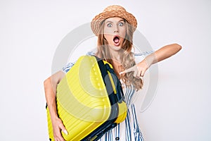 Young caucasian woman with blond hair wearing summer dress and holding cabin bag afraid and shocked with surprise and amazed