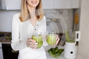 Young caucasian woman with blonde hair making detox cocktail in blender with fresh green vegetables on kitchen at home