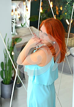 Young Caucasian woman in a blue chemise-dress. A cute beauty with red long hair looks thoughtfully out the window while standing