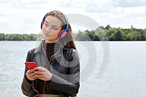 Young caucasian woman in blue red headphones listens to music and typing on the smartphone on the river bank