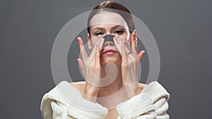 Young caucasian woman cleaning the skin using nose strips from blackheads or black dots isolated on a gray background