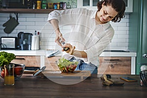 Young Caucasian woman cooking salad from green fresh vegetables while standing in the kitchen at home