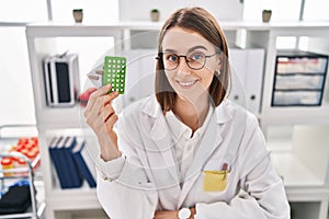 Young caucasian woman doctor holding birth control pills sitting on table at clinic