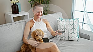 Young caucasian woman with dog watching tv sitting on sofa smiling at home