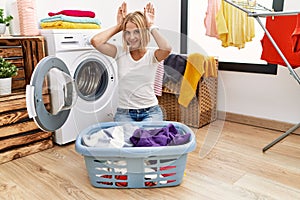 Young caucasian woman doing laundry with clothes in the basket doing bunny ears gesture with hands palms looking cynical and