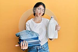 Young caucasian woman doing laundry holding detergent bottle and folded jeans making fish face with mouth and squinting eyes,