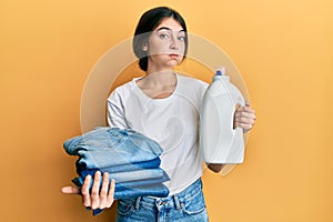 Young caucasian woman doing laundry holding detergent bottle and folded jeans puffing cheeks with funny face