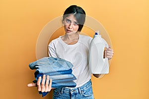 Young caucasian woman doing laundry holding detergent bottle and folded jeans in shock face, looking skeptical and sarcastic,