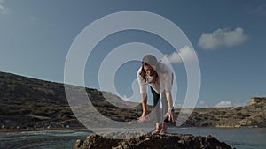 Young caucasian woman is doing yoga breathing and stretching exercises on rock on the beach