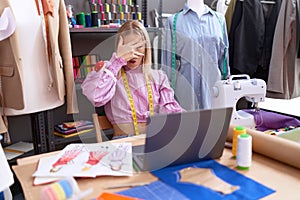 Young caucasian woman dressmaker designer using laptop peeking in shock covering face and eyes with hand, looking through fingers