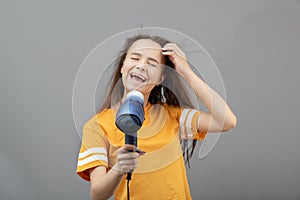 Young caucasian woman dries hair with a hairdryer on a gray background, has fun, sings like a microphone