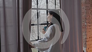 Young caucasian woman drinking morning coffee while looking through the window in winter