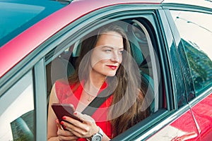 Young caucasian woman driver in red suit holding her cell smart phone and looking away while sitting behind the wheel red car.