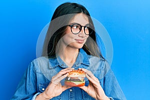 Young caucasian woman eating a tasty classic burger smiling looking to the side and staring away thinking