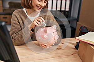 Young caucasian woman ecommerce business worker insert dollar on piggy bank at office