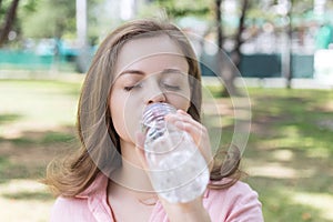 Young caucasian woman girl drinking water from a plastic bottle in green park, close up