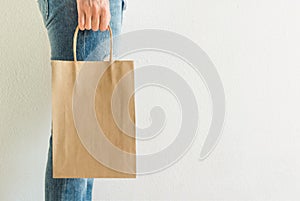 Young caucasian woman girl in jeans holds in hand empty blank mock up brown craft paper bag white wall. Christmas New Year