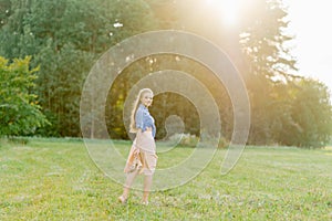 Young Caucasian woman or girl in a summer dress and a denim shirt is walking on the grass, holding shoes in her hands, relaxing