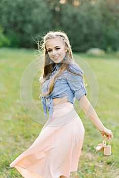 Young Caucasian woman or girl in a summer dress and a denim shirt is walking on the grass, holding shoes in her hands, relaxing