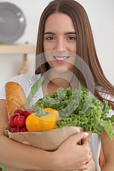 Young caucasian woman in a green apron is holding paper bag full of vegetables and fruits while smiling in kitchen