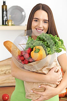 Young caucasian woman in a green apron is holding paper bag full of vegetables and fruits while smiling in kitchen