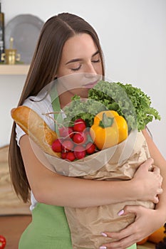 Young caucasian woman in a green apron is holding paper bag full of vegetables and fruits while smiling in kitchen