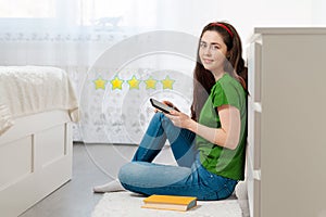 A young Caucasian woman in a green t-shirt sits on the floor and writes a review of a book in her smartphone app. Bright room in