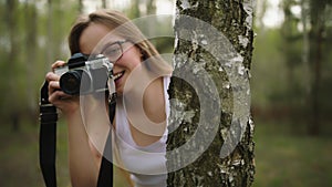 Young caucasian woman hiding behind the tree with a vintage camera. Happy girl photographing the nature