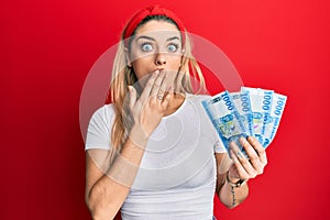 Young caucasian woman holding 1000 hungarian forint banknotes covering mouth with hand, shocked and afraid for mistake
