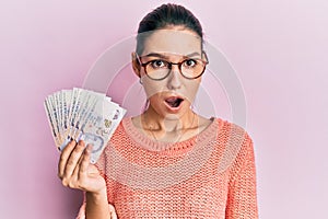 Young caucasian woman holding 2 singapore dollars banknotes scared and amazed with open mouth for surprise, disbelief face