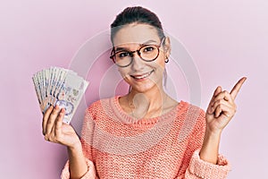 Young caucasian woman holding 2 singapore dollars banknotes smiling happy pointing with hand and finger to the side