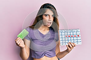 Young caucasian woman holding birth control pills and menstrual calendar in shock face, looking skeptical and sarcastic, surprised